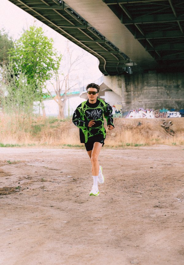 Άνδρας που φοράει αθλητικά ρούχα νέον πράσινου και μαύρου χρώματος και γυαλιά ηλίου τρέχει κάτω από μια τσιμεντένια γέφυρα με γκράφιτι στον τοίχο πίσω του.