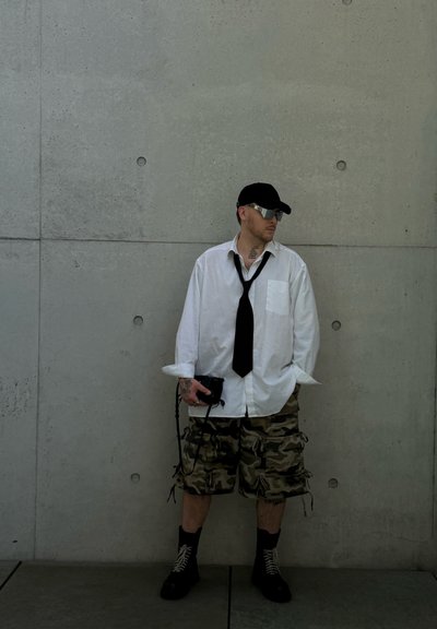 Hombre con camisa blanca, corbata negra, pantalones cortos de camuflaje, gorra negra y gafas de sol, de pie contra una pared de hormigón sosteniendo una bolsa negra.