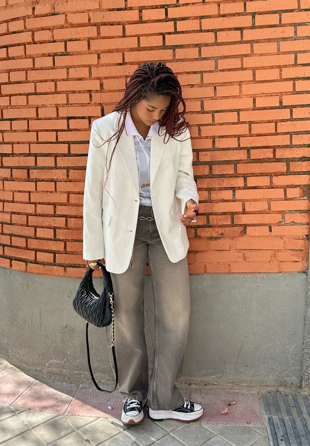 White blazer over a light shirt, grey wide-leg trousers, black handbag, and black trainers. Brick wall background. Natural lighting.
