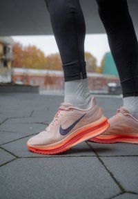 Chaussures de sport Nike en rose clair avec une tige en mesh respirant, un swoosh violet foncé et une semelle amortissante visible orange.