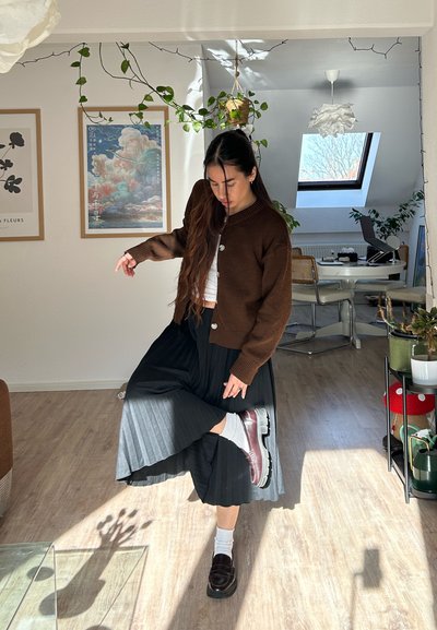 Mujer joven con suéter marrón y falda negra levantando la pierna dentro de una habitación iluminada por el sol, con plantas, obras de arte enmarcadas y una ventana tipo tragaluz.