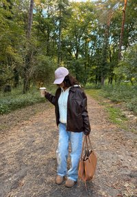 Veste en cuir marron portée sur un pull à mailles crème, jeans bleu clair, chaussons beiges et un sac fourre-tout marron. Tient une tasse de café.