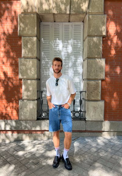 Camisa blanca de manga corta, pantalones cortos de mezclilla, mocasines negros y calcetines con estampado. De pie frente a una pared de ladrillos y una ventana con persianas cerradas.
