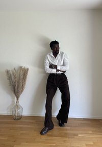 Weißes Langarmhemd, braune weite Hose und dunkle Schuhe. Steht neben einer beigen Vase mit Pampasgras, vor einer strukturierten weißen Wand.