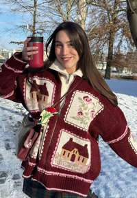 Jeune femme à l'extérieur dans la neige portant un pull bordeaux avec des motifs de maison et floraux, tenant un gobelet rouge, souriante avec de longs cheveux bruns.