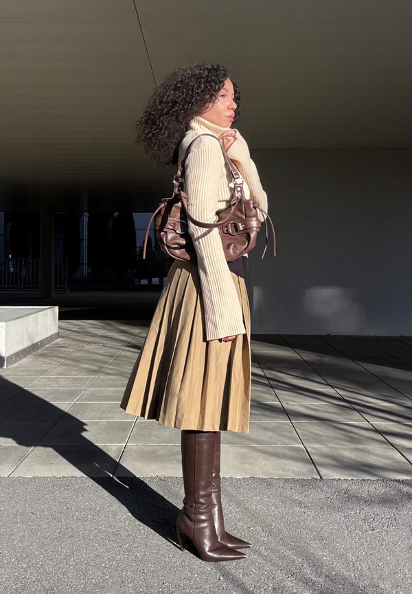 Donna con capelli ricci che indossa un maglione beige a collo alto, gonna plissettata color tan, stivali marroni fino al ginocchio e una borsa marrone, in piedi su un marciapiede piastrellato.