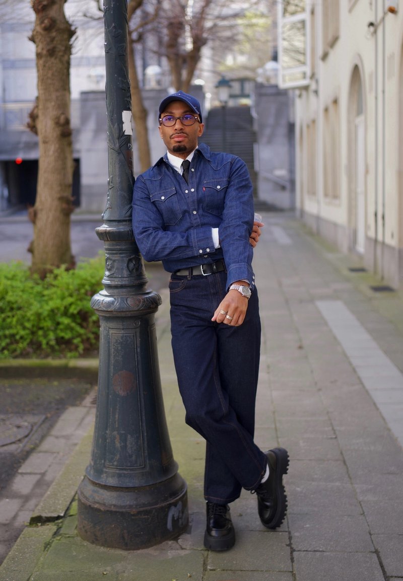 Homme portant une chemise en jean, un jean foncé, une casquette et des lunettes, appuyé contre un lampadaire sur un trottoir urbain, les bras croisés et les jambes croisées.