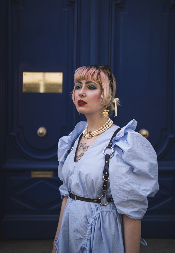 Personne aux cheveux courts roses, maquillage audacieux et rouge à lèvres rouge, portant une robe bleu clair à manches bouffantes et un collier de perles, debout devant une porte bleu foncé.