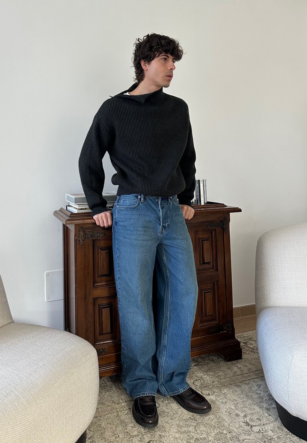 Ung mann som lener seg på et trefatskap, iført svart genser, løse blå jeans og svarte sko, stående mellom beige lenestoler i stuen.