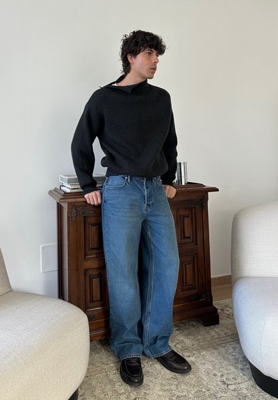 Joven apoyado en un armario de madera, vistiendo un suéter negro, jeans azules holgados y zapatos negros, de pie entre sillones beige en la sala de estar.