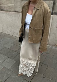 Vrouw in een tan suède jas, witte tanktop, beige satijnen rok met witte kanten zoom, naakte hakken, met een zwarte tas met gouden ketting op het trottoir.
