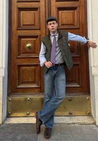 Giovane che indossa un gilet trapuntato verde sopra una camicia azzurro chiaro, berretto grigio, cravatta viola e jeans blu, in piedi contro una porta di legno.