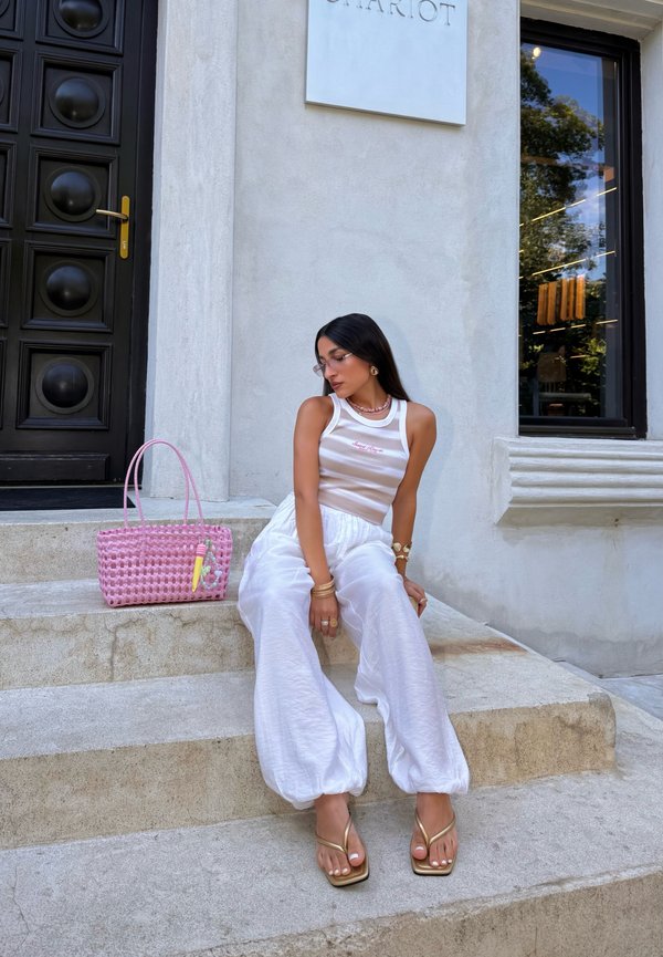 Femme en pantalon blanc et débardeur rayé assise sur des marches en pierre à côté d'un sac à main rose devant un bâtiment moderne avec une porte et une fenêtre noires.