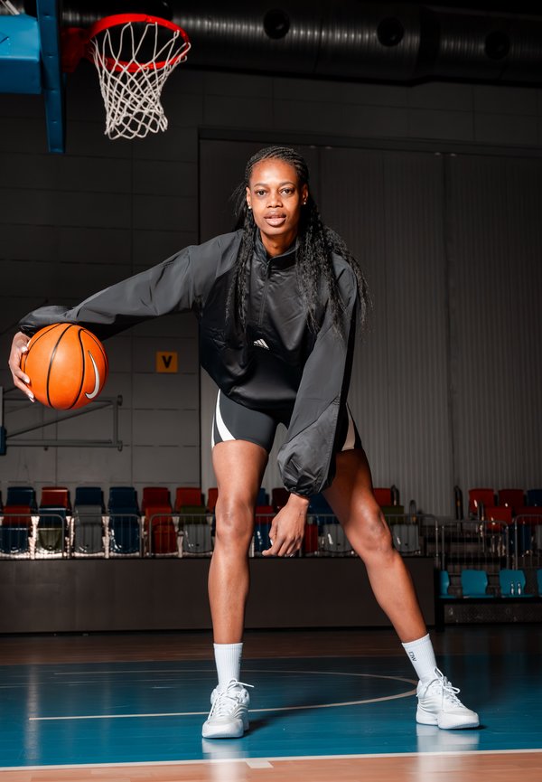 Γυναίκα μπασκετμπολίστρια με μαύρη αθλητική περιβολή ντριμπλάρει μία πορτοκαλί μπάλα σε εσωτερικό γήπεδο κάτω από ένα στεφάνι με κόκκινη στεφάνη και λευκό δίχτυ.