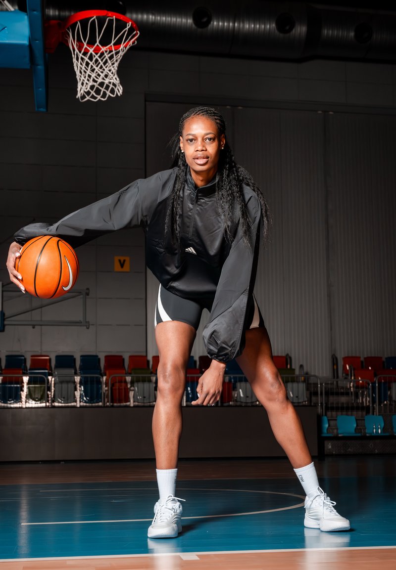 Jugadora de baloncesto femenina con ropa deportiva negra botando un balón naranja en una pista cubierta bajo un aro con borde rojo y red blanca.