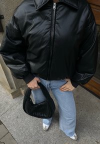 Μαύρο δερμάτινο κοντό μπουφάν με φαρδύ γιακά και μπροστινό φερμουάρ, συνδυασμένο με ανοιχτό μπλε τζιν και μαύρη πλεκτή τσάντα.
