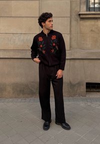 Crna zip-up dukserica s crvenim cvjetnim vezom, u kombinaciji s labavim crnim hlačama i crnim cipelama, stoji naslonjena na kameni zid.