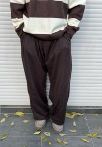 Ruskea ja valkoinen raidallinen ylisuurikokoinen neule nappeineen, yhdistettynä löysiin mustiin housuihin ja harmaisiin saappaisiin teksturoidulla harmaalla pinnalla, jossa on lehtiä.