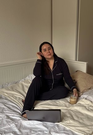 Ensemble de pyjama noir avec des coutures blanches, posé sur un lit avec un ordinateur portable et un verre de boisson glacée, lit léger et oreillers en arrière-plan.