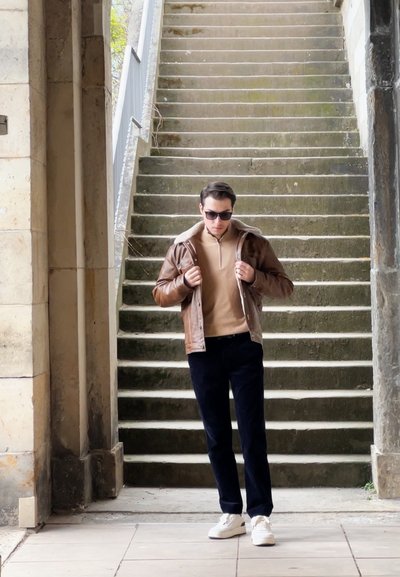 Chaqueta de cuero marrón con cuello de borreguito, suéter beige, pantalones negros y zapatillas blancas, de pie sobre unos escalones de piedra con un fondo texturizado.