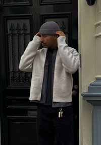 Cardigan gris clair en tricot avec poignets et ourlet côtelés, porté par-dessus une chemise à carreaux foncés, associé à un pantalon noir et un bonnet gris.