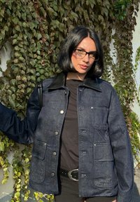 Marineblaue Jeansjacke mit schwarzem Lederkragen, Druckknöpfen und zwei Fronttaschen, über einem schwarzen Hemd getragen, vor grünem Laub Hintergrund.