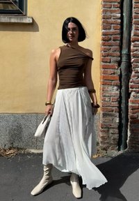 Kobieta z krótkimi czarnymi włosami, ubrana w brązowy top na jedno ramię, białą zwiewną spódnicę, beżowe botki i trzymająca białą torebkę na ulicy w mieście.