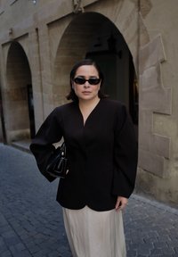 Kobieta w czarnej kurtce, beżowej spódnicy i czarnych okularach przeciwsłonecznych trzymająca czarną torebkę na brukowanej ulicy z kamiennymi budynkami z łukowatymi przejściami.