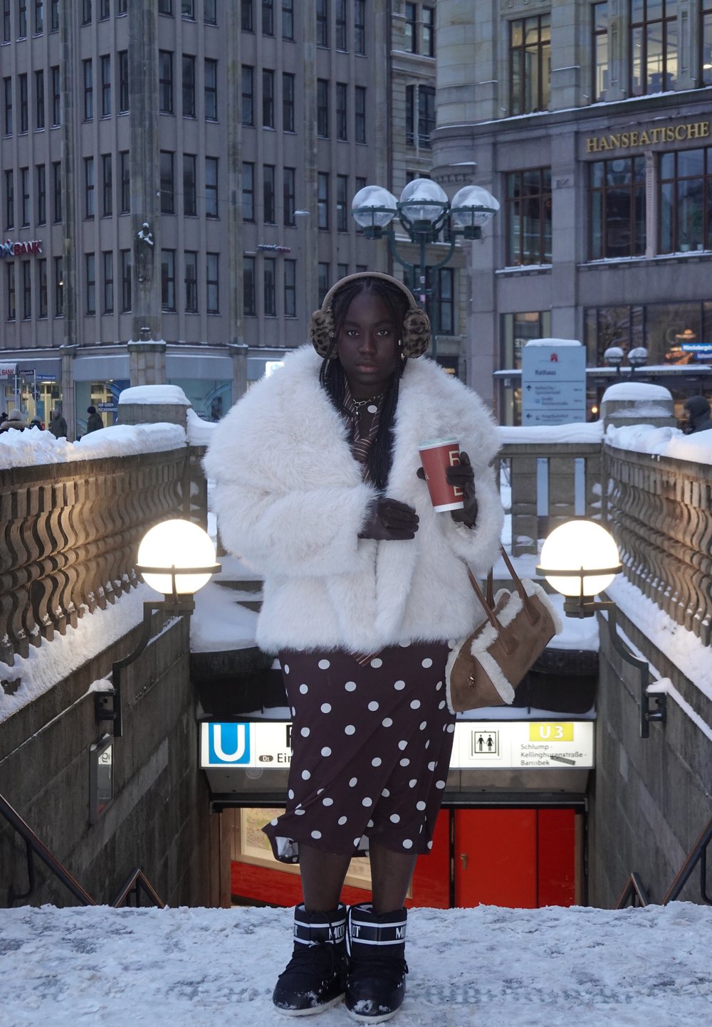 Femme en manteau de fourrure blanc, jupe à pois, cache-oreilles et bottes d'hiver tenant une tasse de café sur des escaliers enneigés menant à une station de métro.