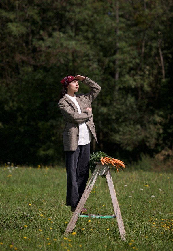 Woman wearing a red bandana and blazer stands on a wooden ladder in a grassy field, with fresh carrots placed on the ladder.