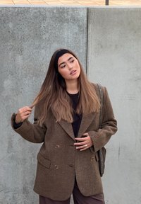 Blazer oversize marrone con trama, con bottoni doppiopetto, indossato su una maglia nera, abbinato a pantaloni scuri e una borsa leggera da spalla.