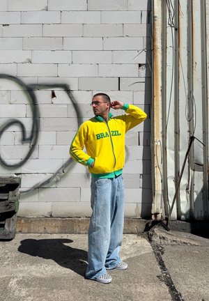 Chaqueta amarilla brillante con cremallera y cuello verde, acompañada de pantalones anchos de mezclilla azul claro y zapatos de cuadros tipo slip-on.