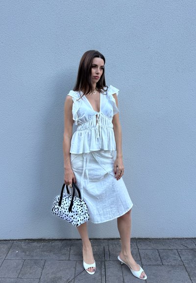 Mujer con blusa blanca sin mangas y falda, sosteniendo un bolso negro con manchas blancas, de pie junto a una pared gris sobre suelo pavimentado.