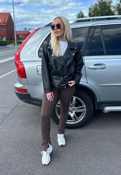 Chaqueta de motorista de cuero negro con herrajes plateados, camiseta blanca, leggins marrones y zapatillas blancas, de pie junto a un SUV plateado.
