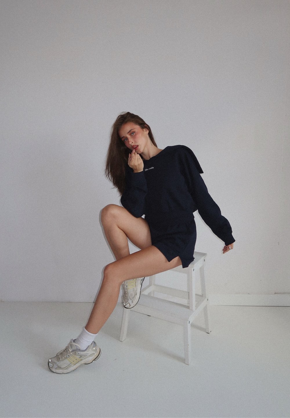 Tengerészkék pulóver válldekorációkkal és logóval, rövidnadrággal párosítva. A modell világosszürke sportcipőt és fehér zoknit visel egy széken.