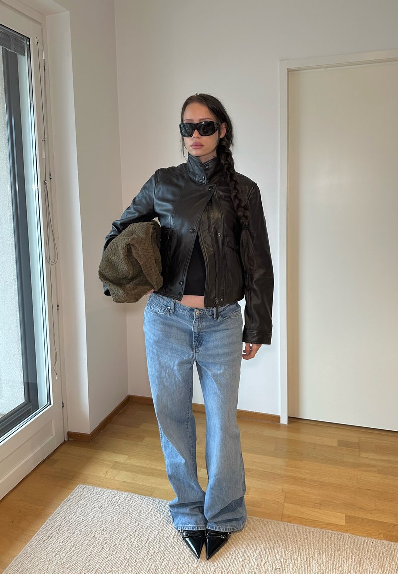 Veste en cuir noir avec boutons-pression, haut noir ajusté, jean flare bleu clair, et escarpins noirs à bout pointu. Cheveux tressés et lunettes de soleil.