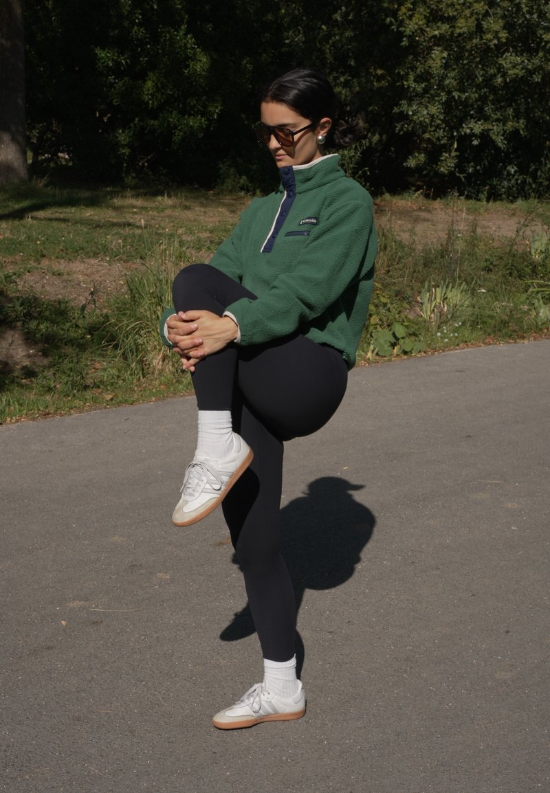 Grüner Fleece-Pullover mit Reißverschluss und Kragen, schwarze Leggings, weiße Socken und Sneakers mit beigen Akzenten. Außenbereich mit Grünpflanzen.