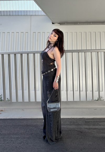 Vestido negro, texturizado, sin mangas, con una abertura lateral y desgastes, combinado con un cinturón metálico, accesorios plateados y un bolso transparente.