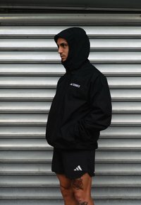Homem de casaco TERREX com capuz preto e calções pretos da Adidas, de pé de lado com as mãos nos bolsos, contra um fundo de persiana metálica.
