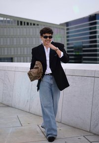 Μαύρη oversized σακάκι πάνω από λευκό πουκάμισο με βολάν, ανοιχτό μπλε παντελόνι σε φαρδύ στιλ και στρογγυλά μαύρα γυαλιά ηλίου. Κρατάει μια μπεζ τσάντα και στέκεται σε εξωτερικό χώρο.
