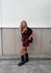 Schwarzes Kleid mit rotem Blumenmuster, taillierte Form, dünne Träger und seitliche Schleife. Schwarze kniehohe Stiefel mit Schnallen. Kombiniert mit einem schwarzen Schal.