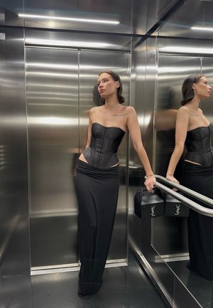 Top corsé negro sin tirantes con costuras detalladas y cortes laterales, combinado con una falda maxi negra ajustada. Sosteniendo dos bolsos negros de YSL.