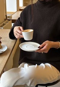 Hvit porselenskopp med gullkant holdt over en matchende skål, foran en koselig kafébakgrunn. Mørk genser og hvite bukser.