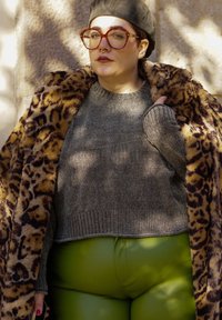 Γκρι πλεχτό πουλόβερ με ριμπ τελείωμα, παλτό από τεχνητό γούνινο λεοπάρδαλη και πράσινο δερμάτινο παντελόνι με γυαλιστερή υφή. Τονισμένο από μεγάλα γυαλιά.