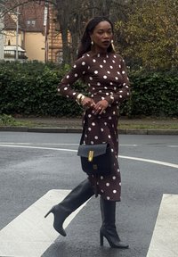 Robe marron à pois avec une silhouette ajustée, associée à des bottes noires montant jusqu'au genou et un sac à main noir structuré avec un accent doré.
