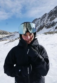 Schwarze Skijacke mit hohem Kragen, schwarze Handschuhe und verspiegelte Skibrille. Im Hintergrund eine verschneite Landschaft und felsige Berge.