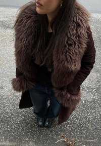 Femme portant un manteau marron foncé à col en fourrure, une chemise noire, un jean bleu et des chaussures noires à bout carré, debout sur un trottoir moucheté.