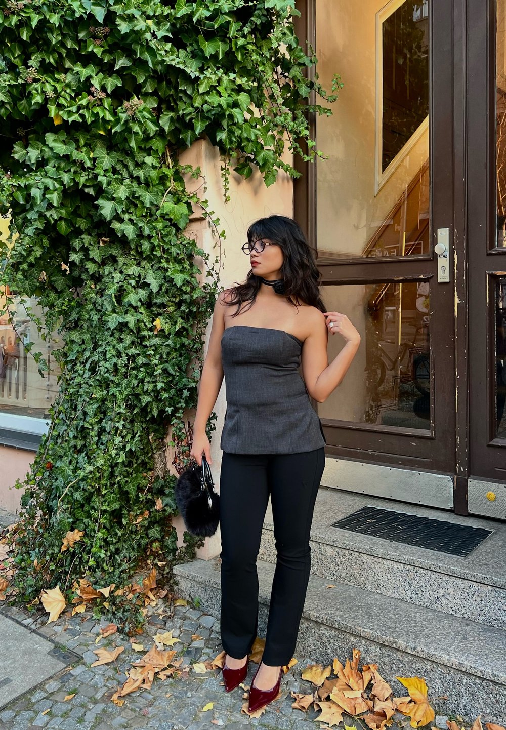 Dark grey strapless top with a fitted design, black skinny trousers, and burgundy high heels. Accessorised with a black faux fur clutch. Green ivy background.