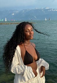 Svart bikiniöverdel, vit lös skjorta, vågigt svart hår. Vatten med båtar i bakgrunden, berg i fjärran. Mjukt ljus.