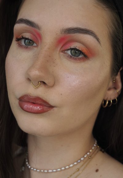 Mujer joven con audaz sombra de ojos en tonos naranja y rojo, labios marrones brillantes, piercing en el septum, múltiples perforaciones en las orejas y collares en capas con perlas y cuentas doradas.